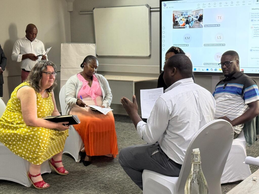 African and European men and women sit facing each other. A woman in yellow is talking. People stand around them listening